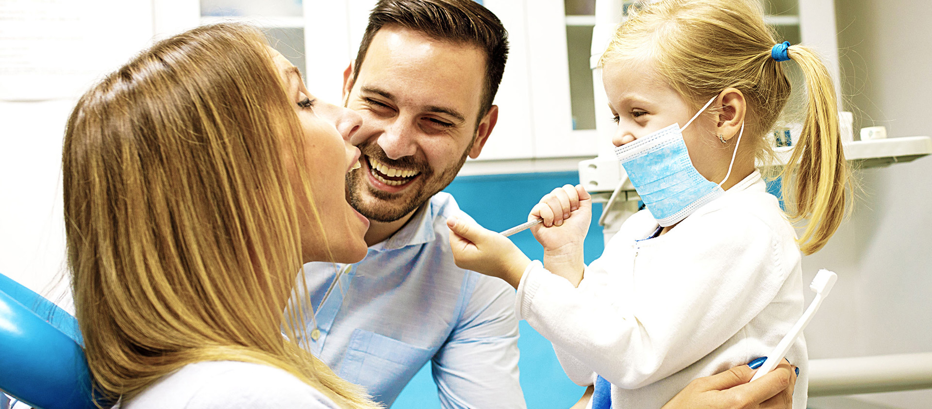 Child examining mother's teeth while father looks on laughing
